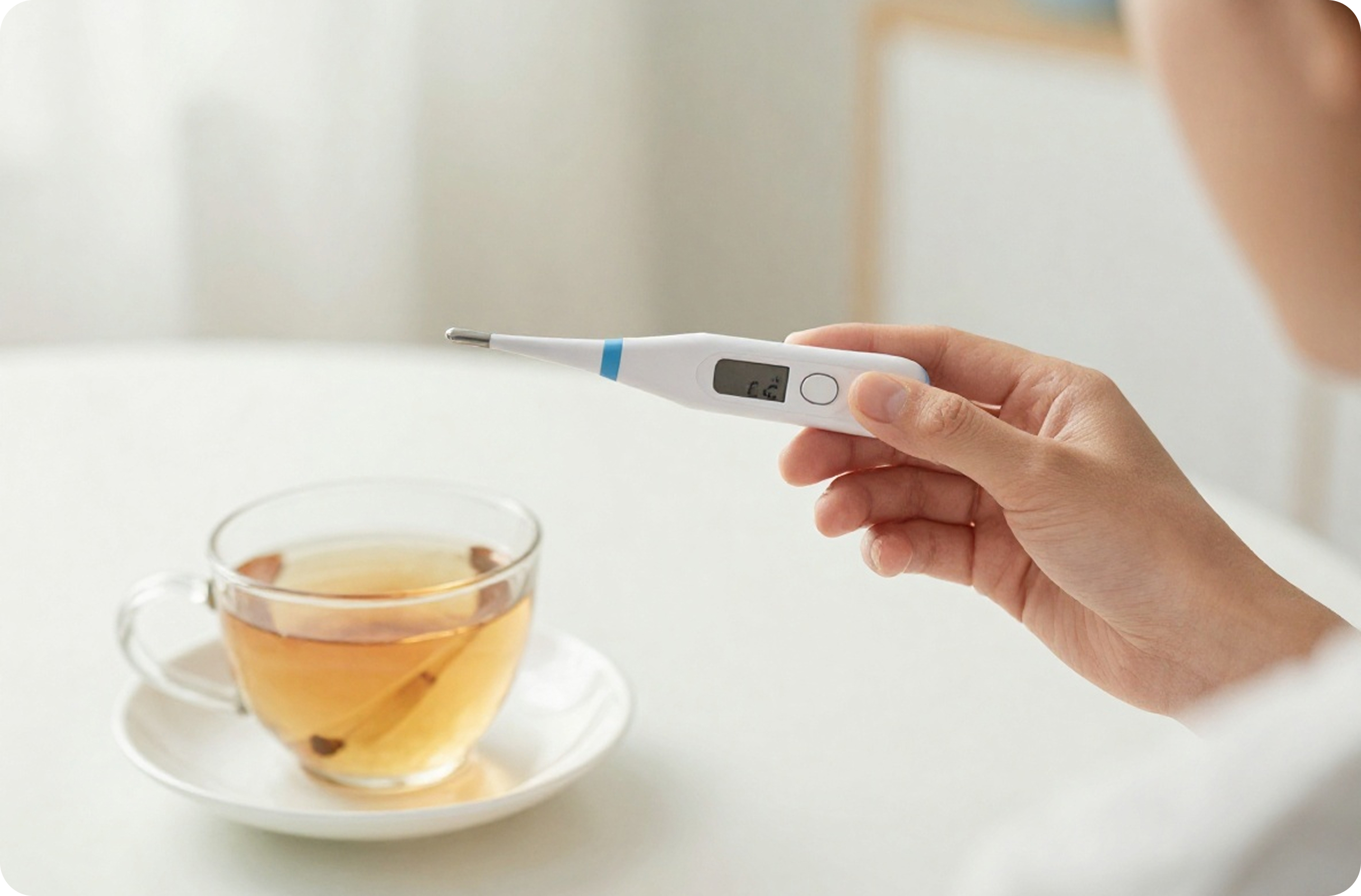 Hand holding a digital thermometer above a cup of tea on a white table